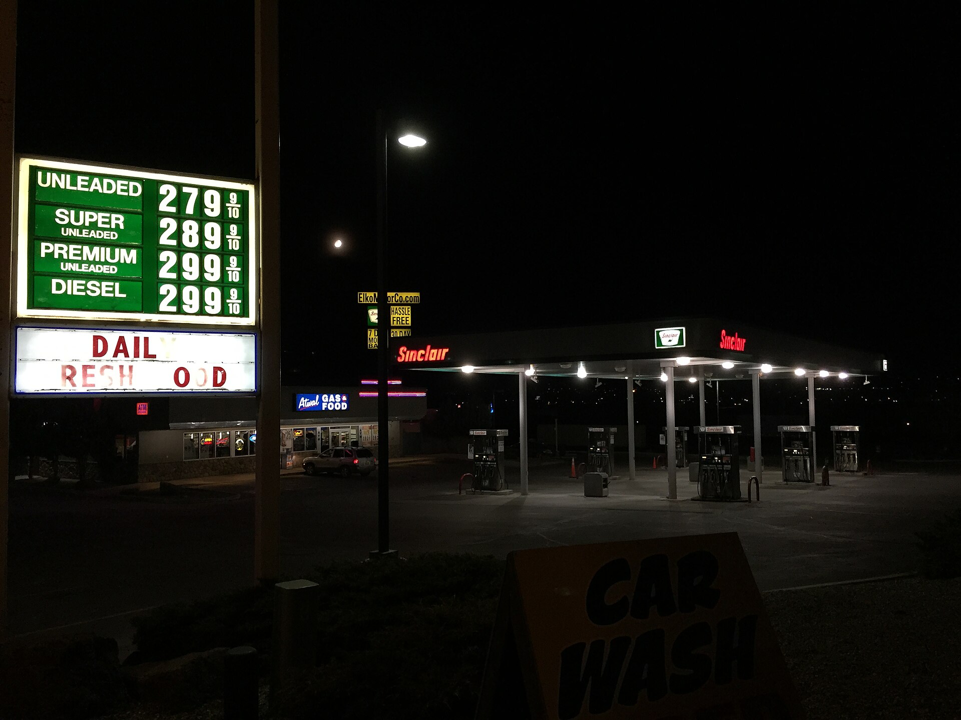 An empty, lit gas station forecourt at night: a price sign reading 279.9 unleaded, a canopy over six pumps, one parked SUV outside the store, the moon above
