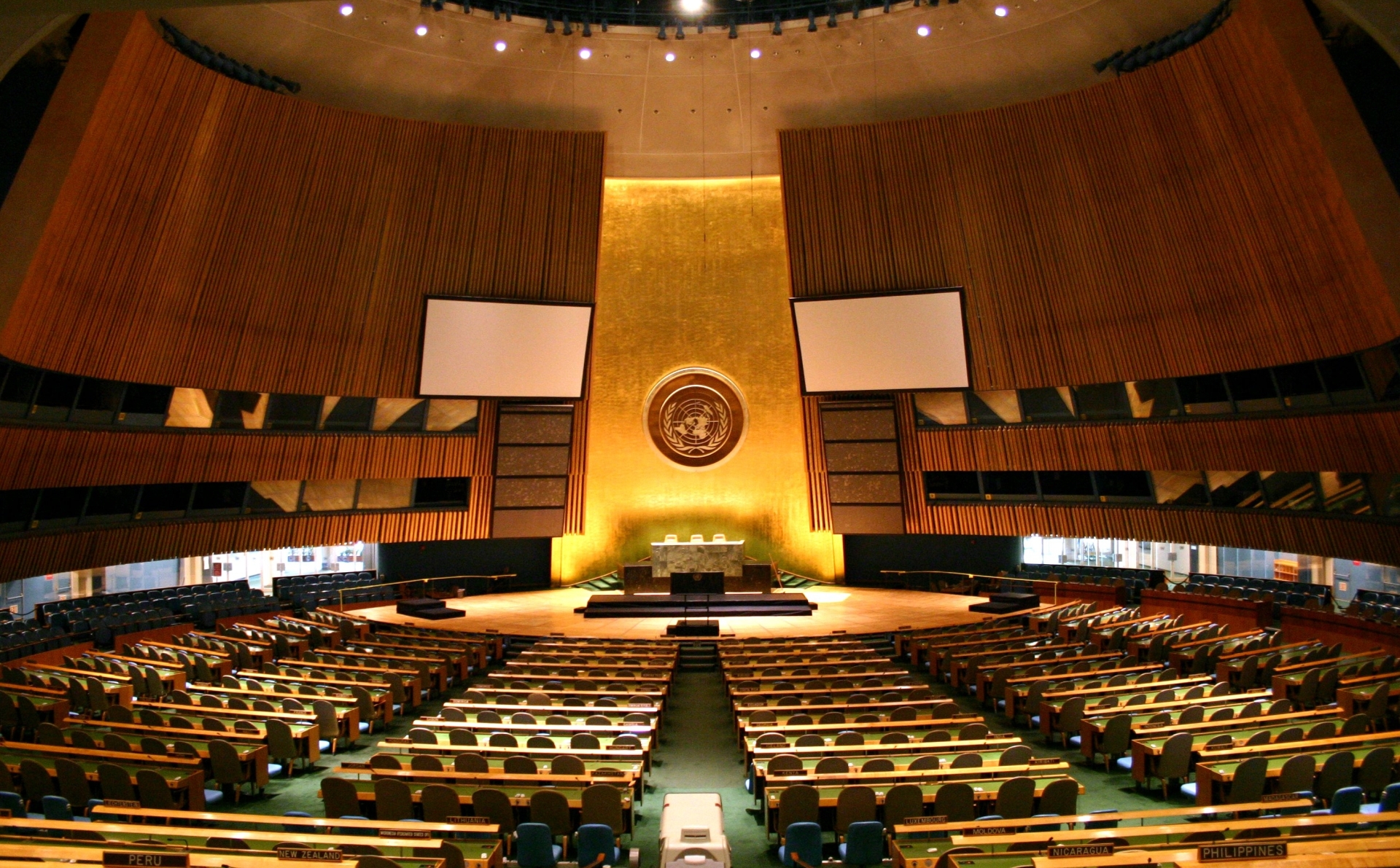The empty United Nations General Assembly hall photographed from the back of the chamber: curved rows of pale wooden desks with green seats descending toward a raised marble rostrum, the gold-lit wall behind it carrying the circular United Nations emblem, two blank projection screens on either side, ribbed wood panelling curving up to a domed ceiling, and small country nameplates on the nearest desks reading Peru, New Zealand, Lithuania, Luxembourg, Moldova, Nicaragua and Philippines; no person is visible