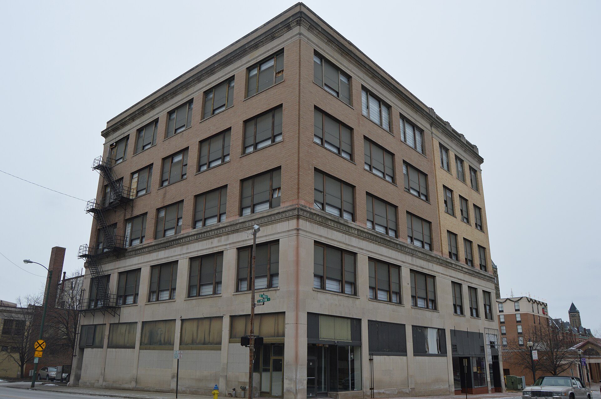 A street corner in downtown Springfield, Ohio, photographed from across the intersection on an overcast winter day. A five-storey brick-and-stone commercial block fills most of the frame, its ground-floor shopfronts boarded and shuttered, a black iron fire escape zigzagging down the left-hand wall. Green street blades on a pole at the corner read E HIGH ST and S LIMESTONE ST. A yellow railroad crossing sign and two parking signs stand on the near pavement. Bare trees and a parked car are at the edges. There is nobody in the picture.