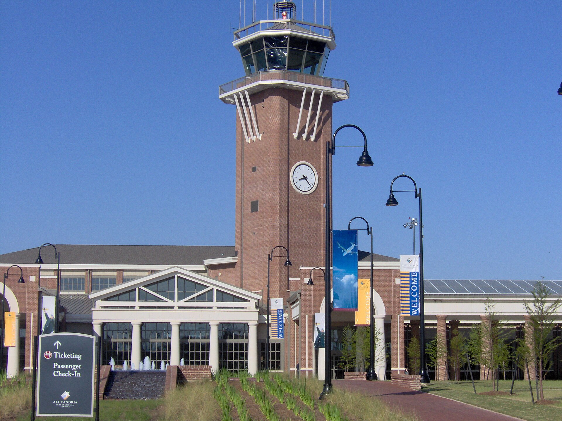 A brick airport terminal under a clear blue sky with a glass-topped control tower and a clock above the entrance, banners reading WELCOME along the walkway, and a sign in the foreground reading Ticketing, Passenger Check-In, Alexandria International Airport; nobody in the frame