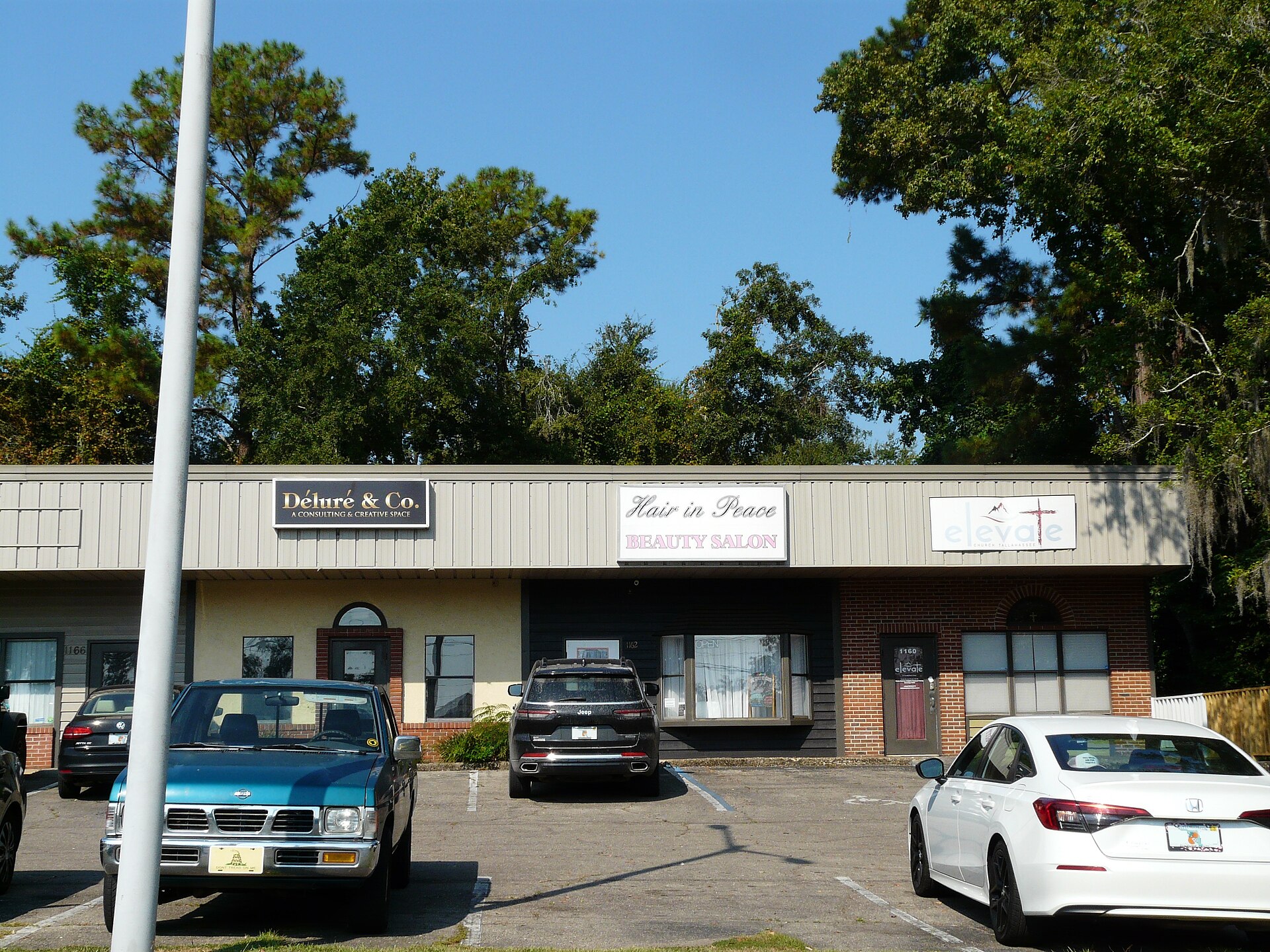 A one-storey strip-centre building under a clear blue sky, photographed from its parking lot: three lit sign boards along the roofline read 'Déluré & Co. A Consulting & Creative Space', 'Hair in Peace Beauty Salon' and 'elevate Church Tallahassee', with unit numbers 1166, 1162 and 1160 beside three separate doors, an OPEN sign in the salon's curtained window, pine and live oak trees behind the roof, and four parked cars with Florida plates in the foreground; no person is visible