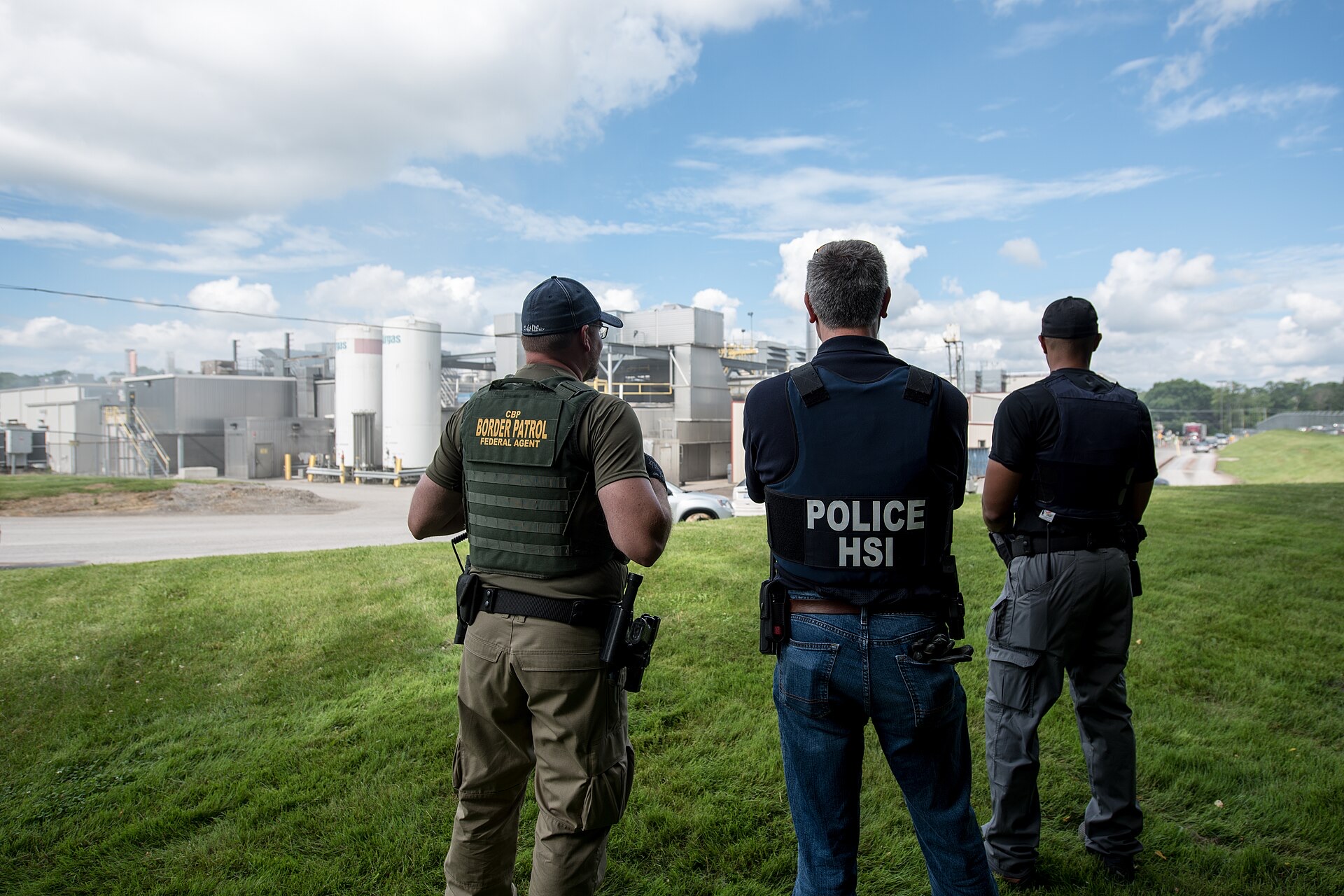 Three federal agents in CBP Border Patrol and HSI Police vests stand with their backs to the camera facing a meat-processing plant in Salem, Ohio