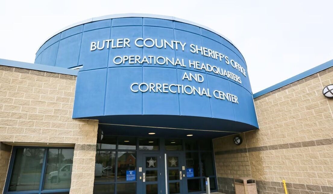 The front entrance of a low public building photographed from close in and slightly below. A curved blue metal canopy wraps across the upper half of the frame, carrying raised silver letters that read BUTLER COUNTY SHERIFF'S OFFICE, OPERATIONAL HEADQUARTERS AND CORRECTIONAL CENTER. Below it, beige split-face stone walls flank a recessed entrance with dark-framed glass doors, each door marked with a small silver sheriff's star. Small blue notice placards are fixed beside the doors, a round light fixture is mounted on the stone to the right, and a metal waste bin stands on the pavement. The sky behind is flat and white. There is nobody in the picture.