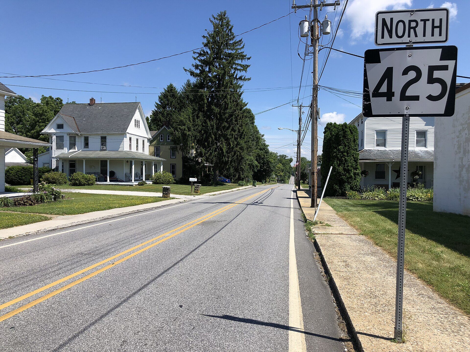 An empty two-lane road running away from the camera through a small town, photographed from the shoulder. A double yellow line runs down the middle and a solid white fog line marks the right edge, with faint tyre marks curving across the asphalt. On the right, close to the lens, a metal signpost carries a small white NORTH plate above a Pennsylvania keystone-shaped route marker reading 425. Beyond it are a grass verge, a concrete footpath, a wooden utility pole strung with wires, a clipped evergreen hedge and white clapboard houses with covered porches. On the left are more white and pale-green houses with porches, a tall spruce, mown lawns and a mailbox at the kerb. The sky is clear blue. There is no person and no moving vehicle in the picture.