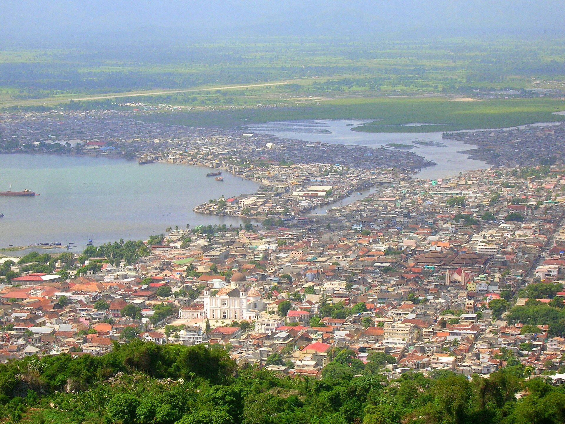 A hillside view over a dense coastal city: tin and tile roofs packed along a harbour, a white twin-towered cathedral with a dark dome near the centre, a river mouth and green plain behind, hills in the haze; nobody identifiable in the frame