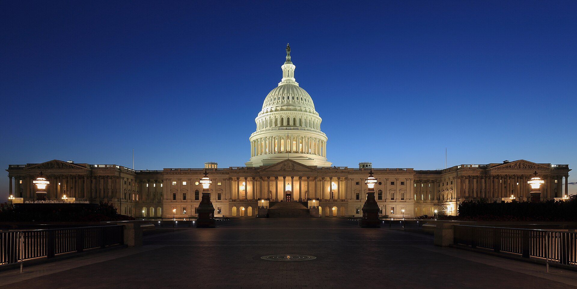 The east front of the United States Capitol at dusk, its dome lit against a deep blue sky, the plaza in front of it empty