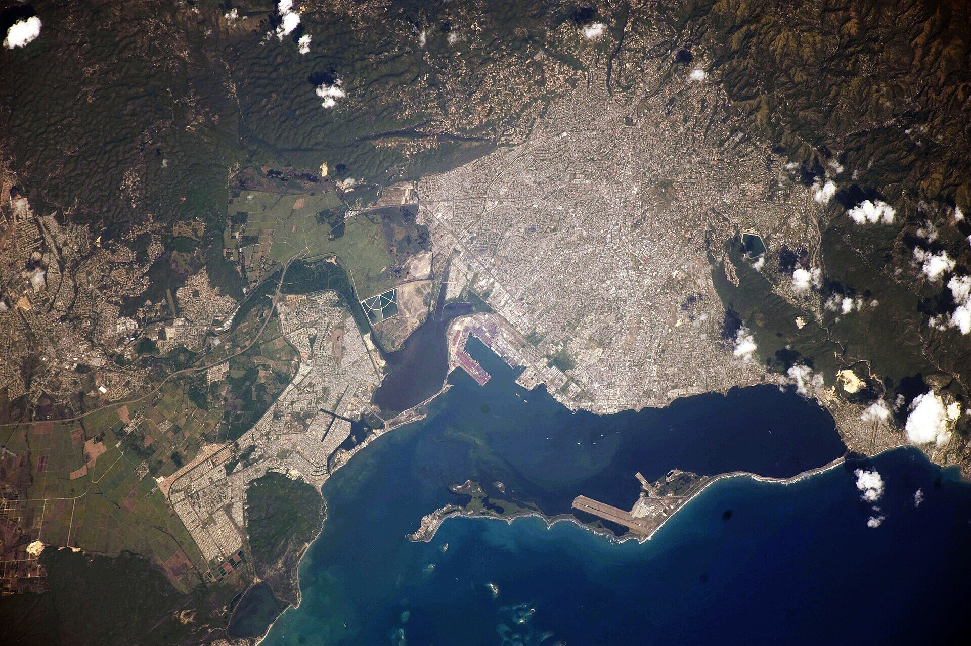 Kingston, Jamaica, photographed from orbit. The city spreads across a coastal plain between green hills and a deep blue harbour, with a long narrow spit of land curving across the bottom of the harbour mouth and a single runway running along it. Scattered white clouds sit over the hills. No people are visible.