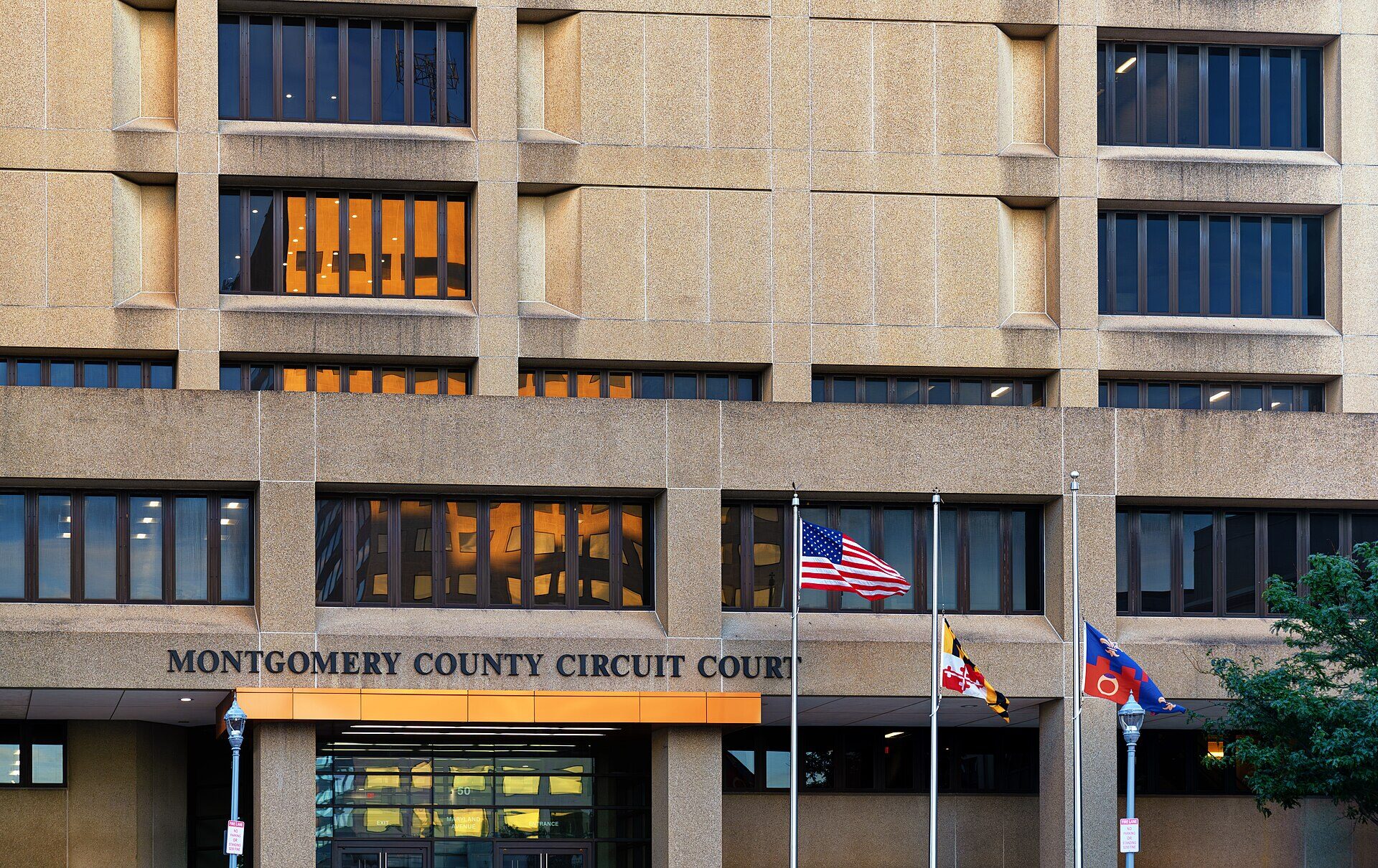The front of a tan concrete government building, several storeys of recessed windows above a low entrance canopy. Carved into the stone above the doors are the words MONTGOMERY COUNTY CIRCUIT COURT. Three flagpoles stand at the right of the entrance flying the United States flag, the Maryland state flag and a county flag. No people are in the frame.