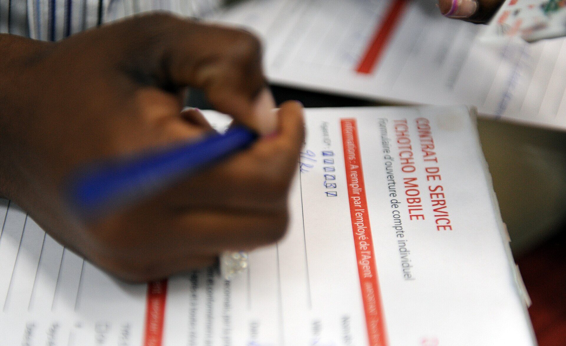 A close photograph of a hand holding a blue ballpoint pen over a printed form on a counter. The form is turned sideways in the frame and its red and blue heading reads CONTRAT DE SERVICE TCHOTCHO MOBILE, with a smaller line beneath it reading Formulaire d'ouverture de compte individuel, and a red bar across it reading Informations a remplir par l'employe de l'Agent. A row of handwritten digits has been entered in small boxes beside the word Agent. More printed forms and papers are stacked underneath and behind. No faces are visible.