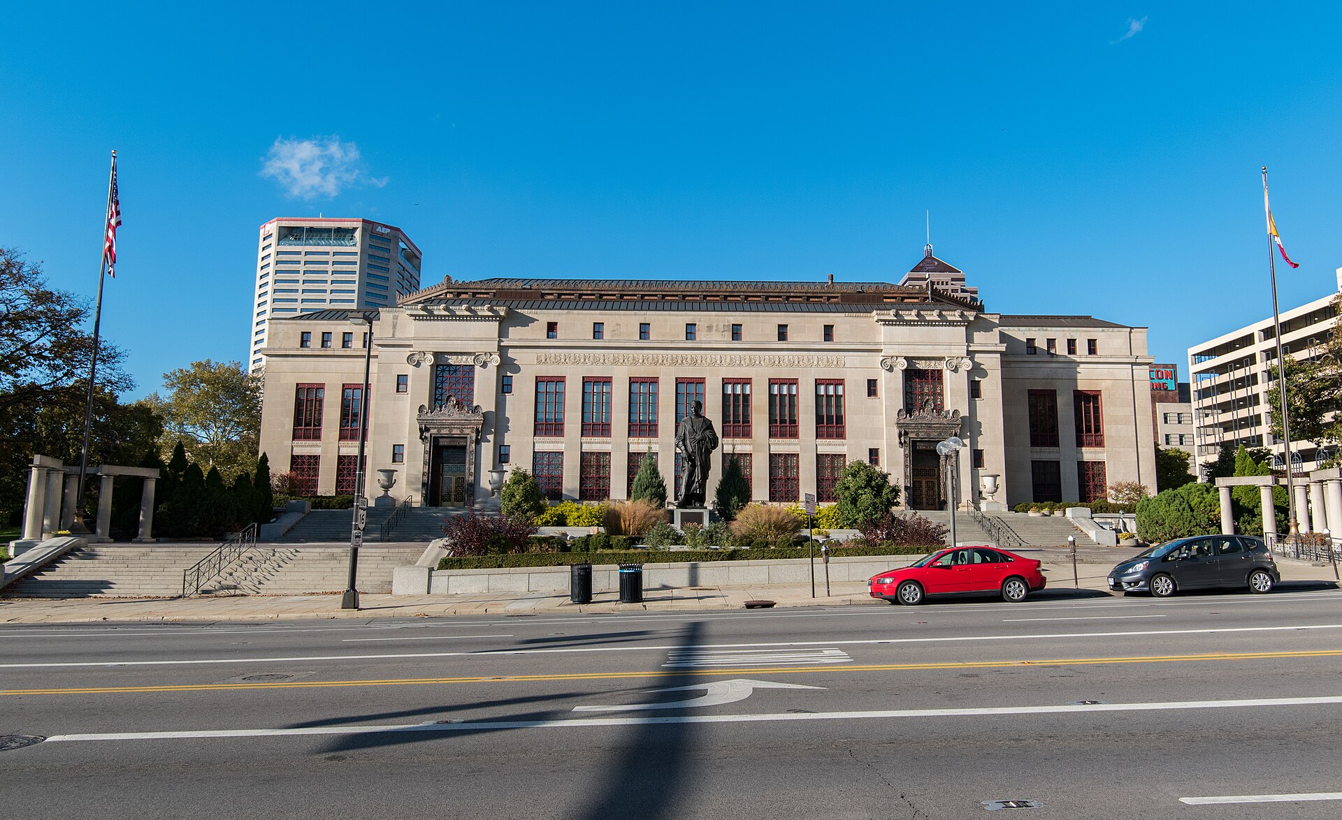 Columbus City Hall photographed from across a wide empty street: a long two-storey limestone building with ionic columns, tall red-framed windows and a low clipped roofline, a bronze statue on a plinth in the garden in front of it, an American flag on a pole at the left and the swallowtail Ohio state flag at the right, the AEP office tower rising behind it against a clear blue sky. Two parked cars at the kerb and no one on the pavement.