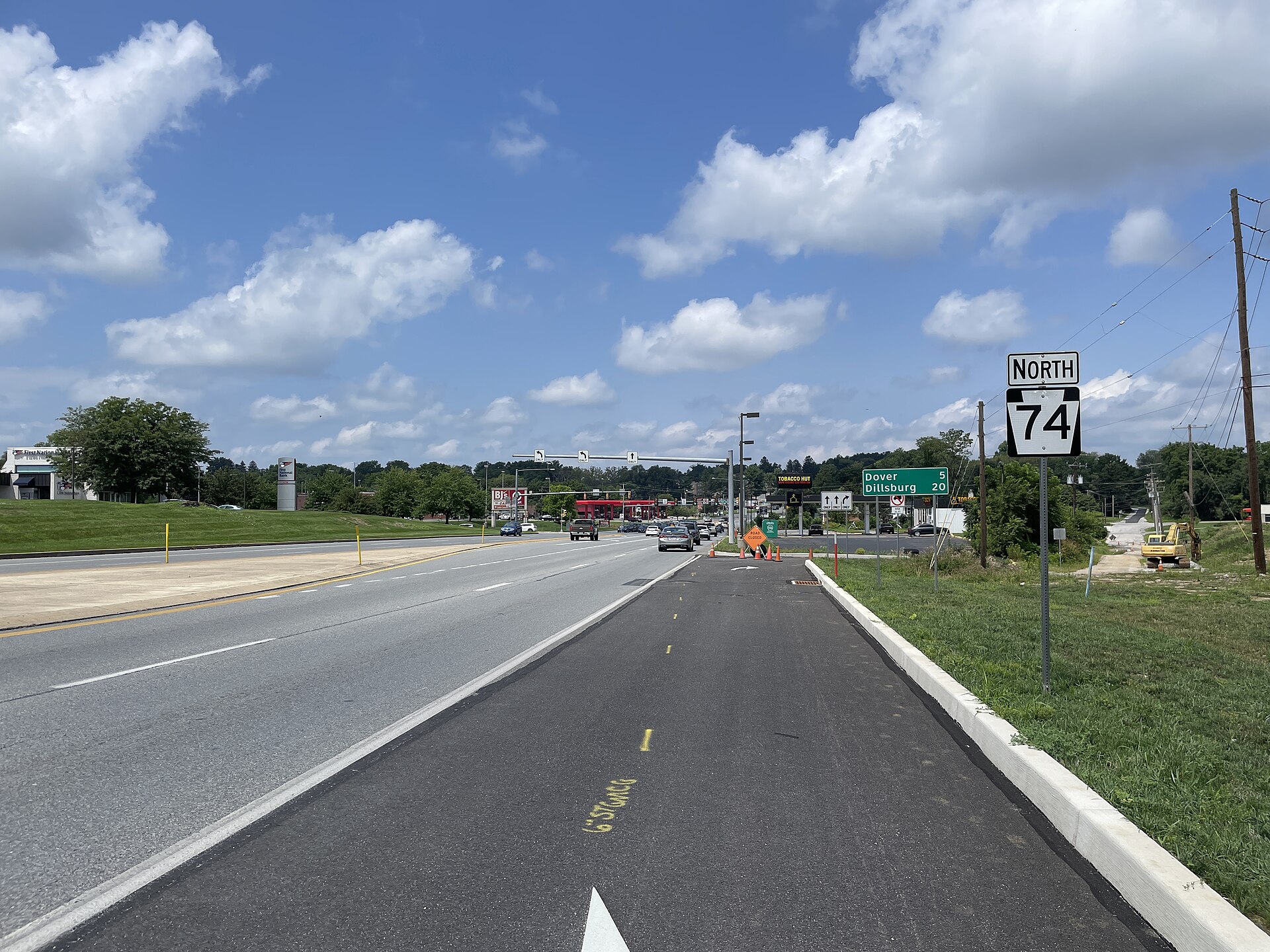 A wide suburban highway under a blue sky with scattered cloud: a freshly paved right-hand lane in the foreground, a concrete kerb and grass verge on the right carrying a black-and-white NORTH 74 Pennsylvania route shield, a green distance sign reading Dover 5 and Dillsburg 20, traffic signals and a line of small cars far ahead, orange cones and a yellow excavator at the roadside; no person is visible or identifiable