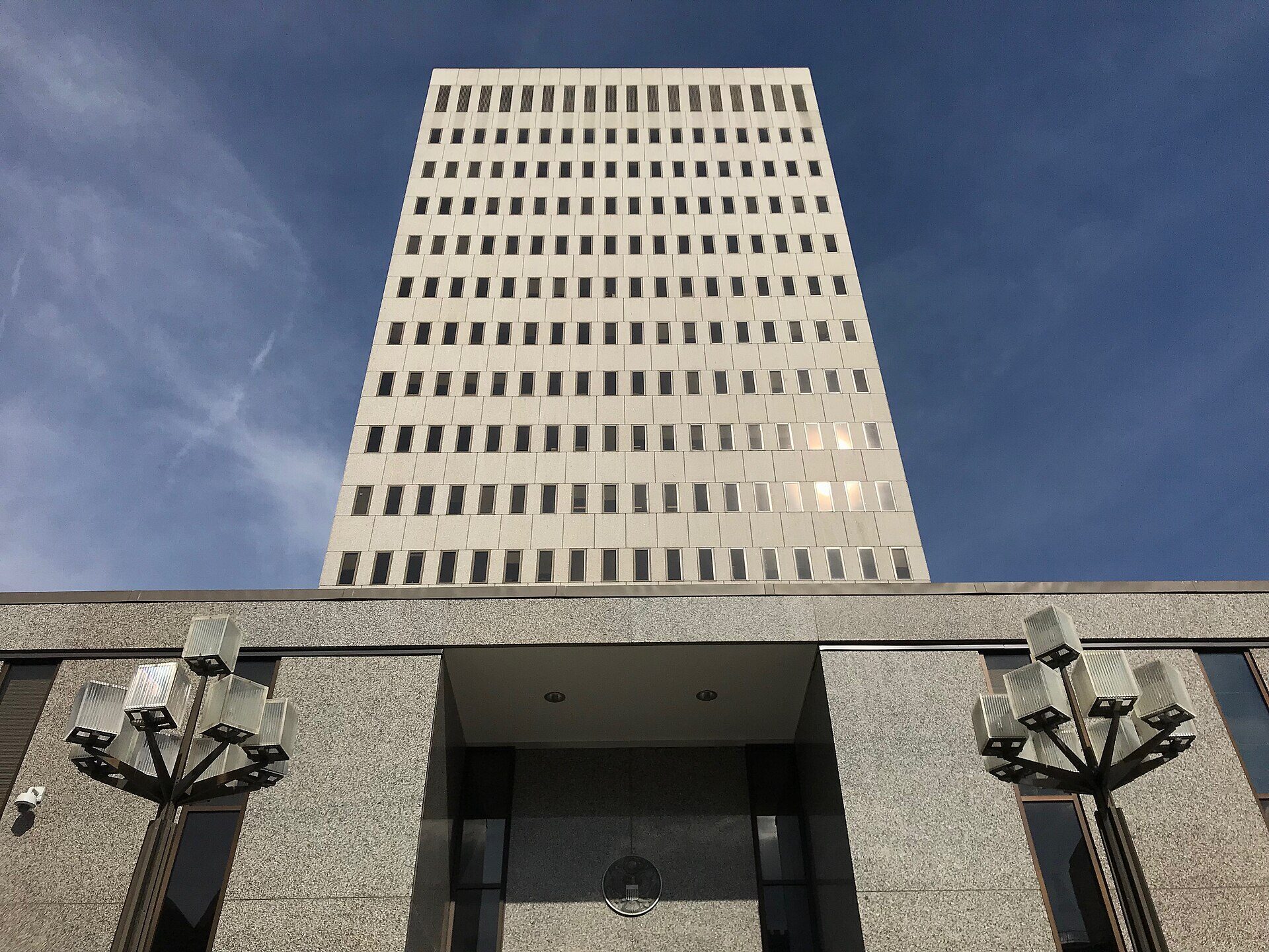Looking almost straight up at a pale, rectangular mid-century federal office tower against a deep blue sky. The tower's long flat face is covered in rows of small, narrow, evenly spaced windows, perhaps fifteen storeys of them, with a few lit from inside. Below it sits a low granite-faced entrance pavilion with a recessed doorway, dark glass panels, and two tall lamp-posts whose clustered square glass lanterns rise on either side. A small round seal hangs above the entrance. The pavement is out of frame and nobody is in the picture.