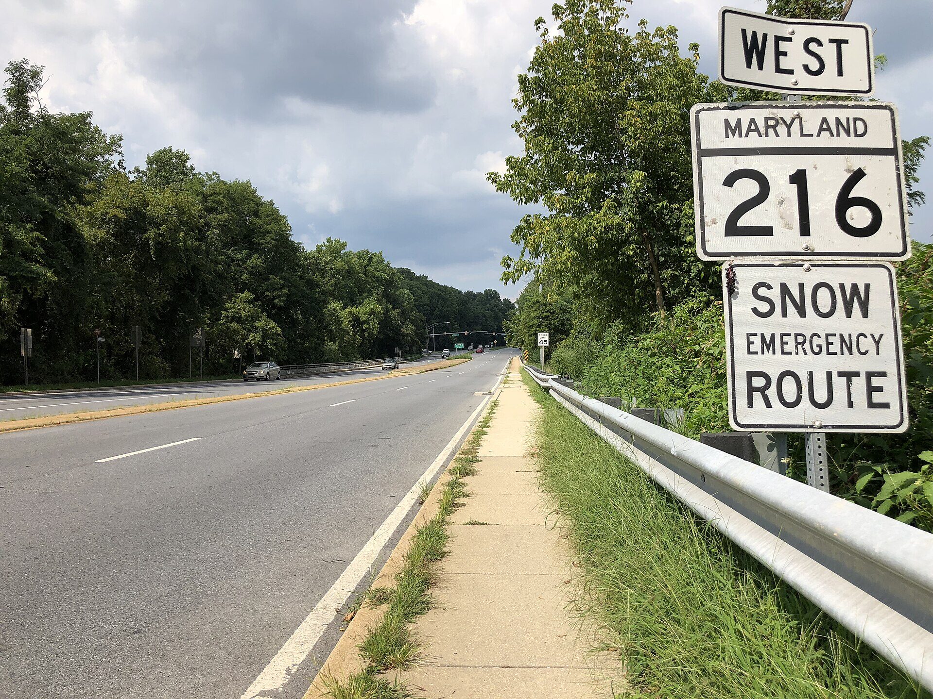 A four-lane road running away from the camera under a grey, clouded sky, with trees on both sides and an empty concrete sidewalk along the right-hand shoulder behind a metal guardrail. Two cars are in the far distance. On the right, close to the camera, a pole carries three white road signs reading WEST, then MARYLAND 216, then SNOW EMERGENCY ROUTE. No person is in the frame.