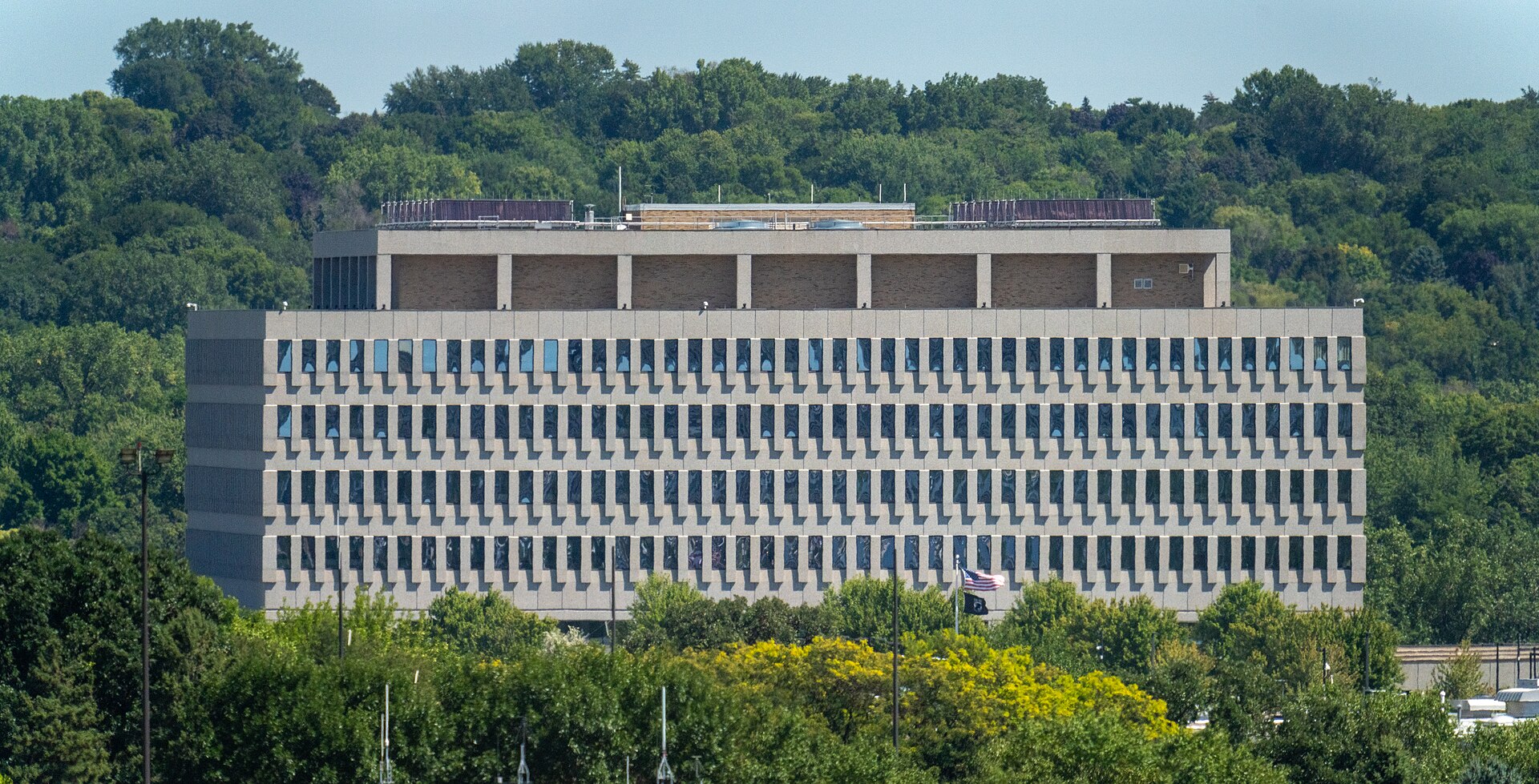 A long concrete-and-glass federal office building on a wooded hillside, an American flag and a POW/MIA flag on poles in front of it, nobody in the frame