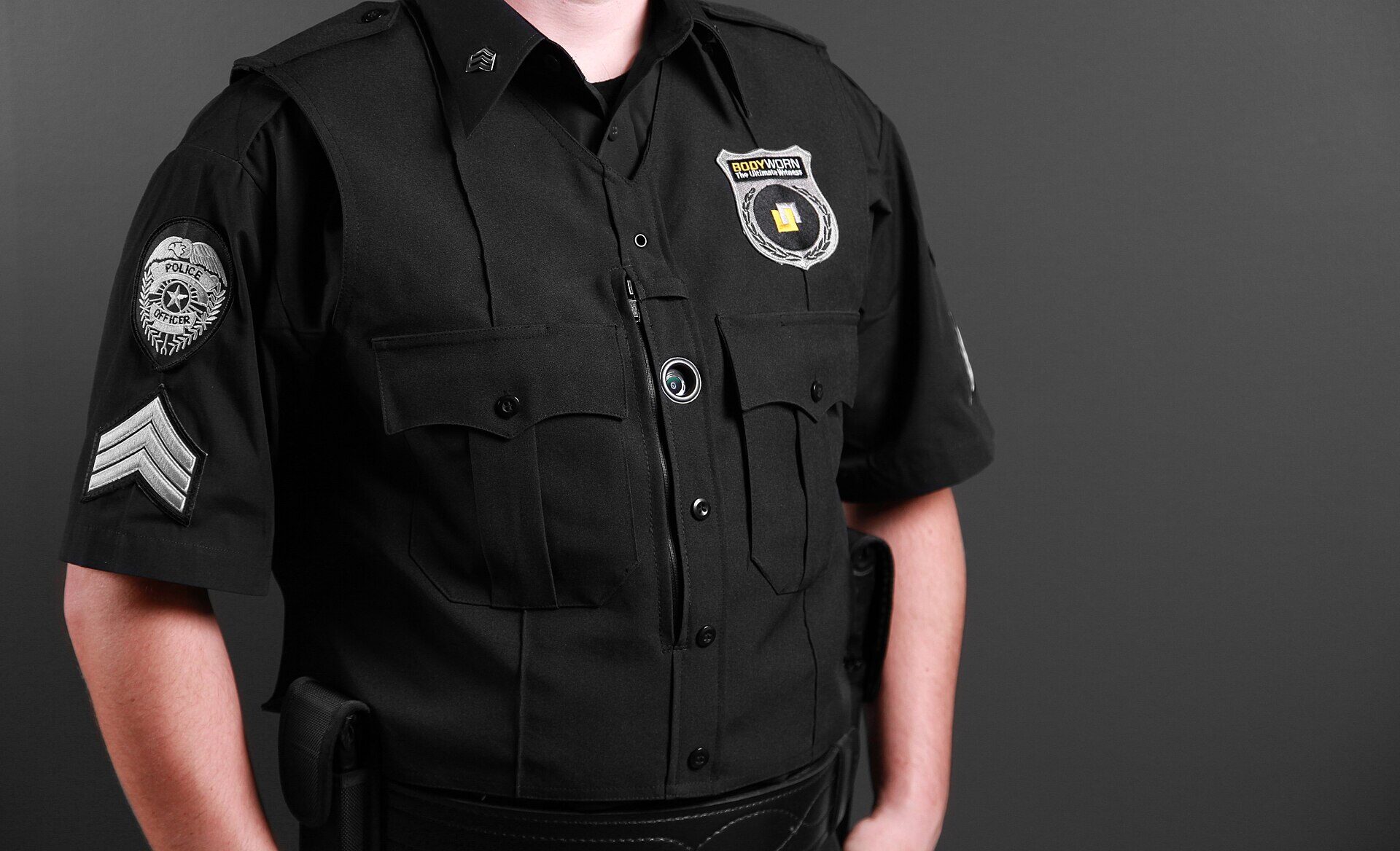 A close-up of the torso of a person in a black municipal police uniform, photographed against a plain grey background. The face is not in the frame. A small round camera lens is fitted on the front of the shirt between the two chest pockets. A round shoulder patch reads POLICE OFFICER and sergeant's chevrons are stitched below it. A badge-shaped maker's logo on the chest reads BODYWORN.