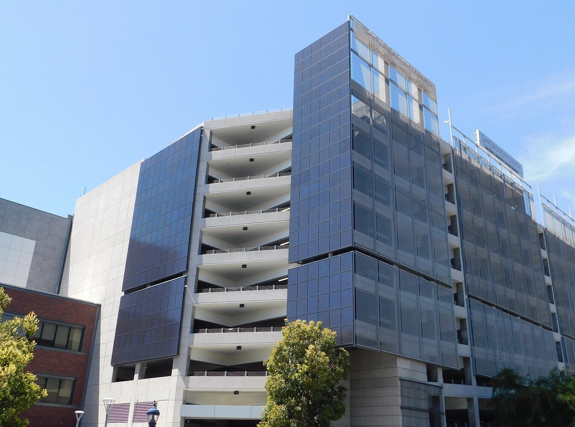 A large concrete parking structure at San Diego City College, photographed from below against a clear blue sky. Dark blue vertical photovoltaic panels cover its corner and side walls, and open parking decks are visible between them. Signs reading SAN DIEGO CITY COLLEGE and SAN DIEGO COMMUNITY COLLEGE DISTRICT are mounted along the top edge. A brick building and two trees stand at the left. No people are in the frame.