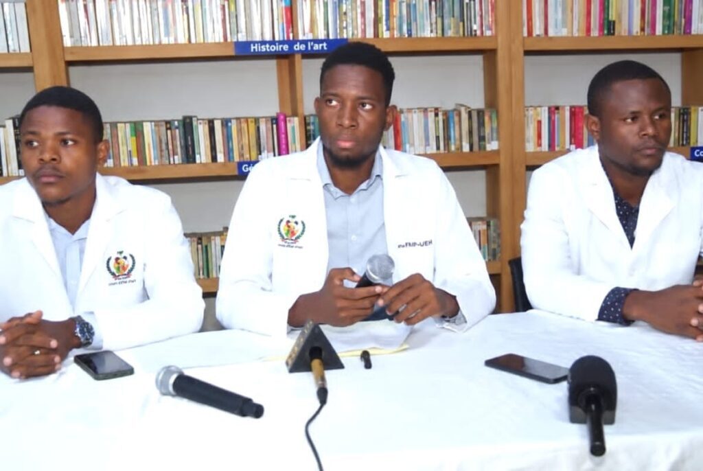 Three medical students in white coats sit behind a long table covered with a white cloth, facing a row of microphones and two voice recorders. Behind them are wooden library shelves filled with books, with French section labels visible above the shelves. One of the three is speaking into a handheld microphone. No patients and no children are in the frame.