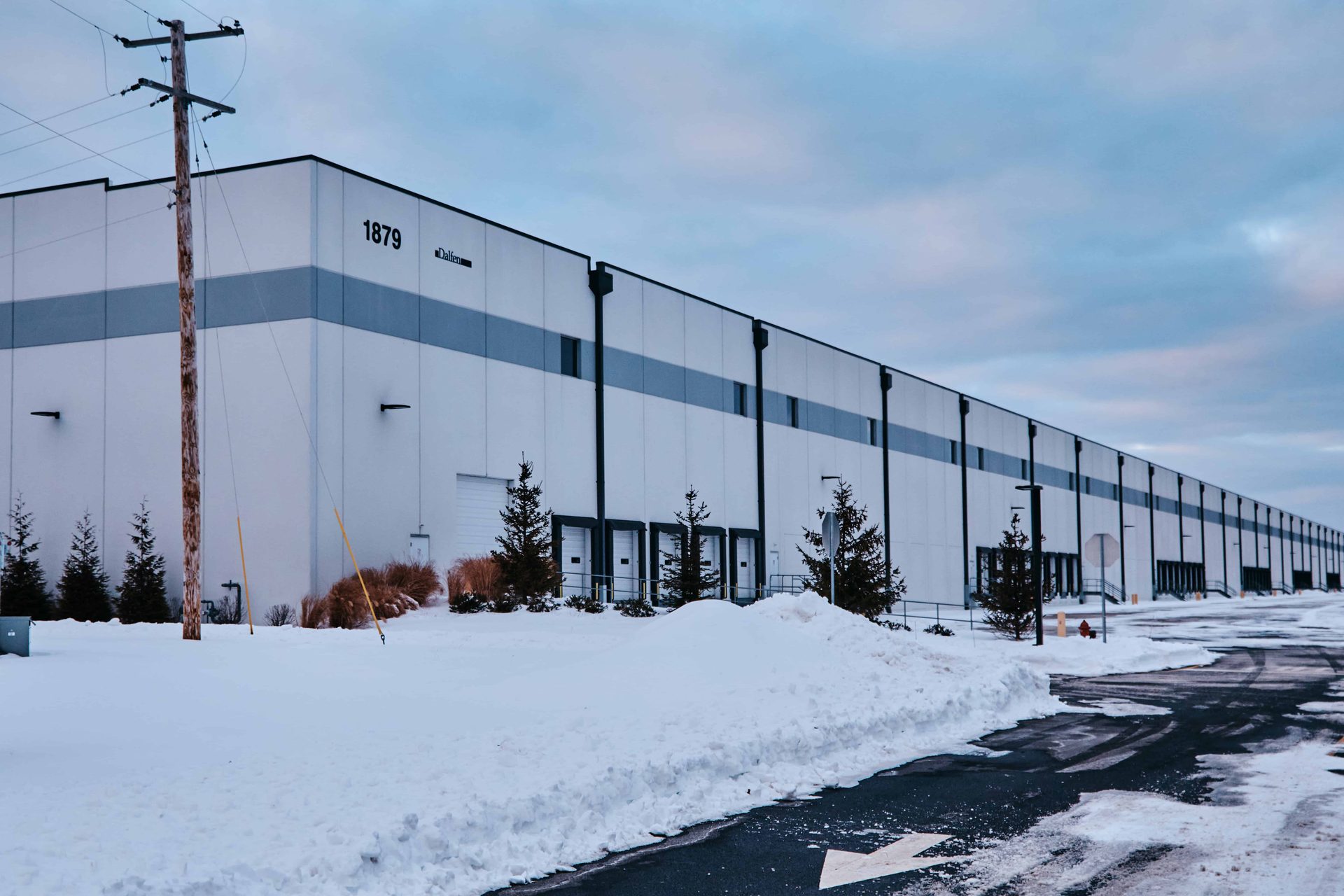 A long white warehouse with a row of loading docks stretching to the horizon, the number 1879 on its corner, snow on the ground and a utility pole in front, nobody in the frame