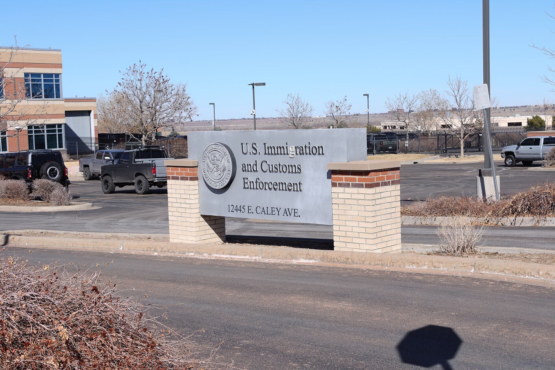 A low grey stone sign on two brick pillars at the entrance to a parking lot, carved with the Department of Homeland Security seal and the words U.S. Immigration and Customs Enforcement above a street address. Parked pickup trucks and a bare-branched tree stand behind it under a clear winter sky. No people are in the frame.