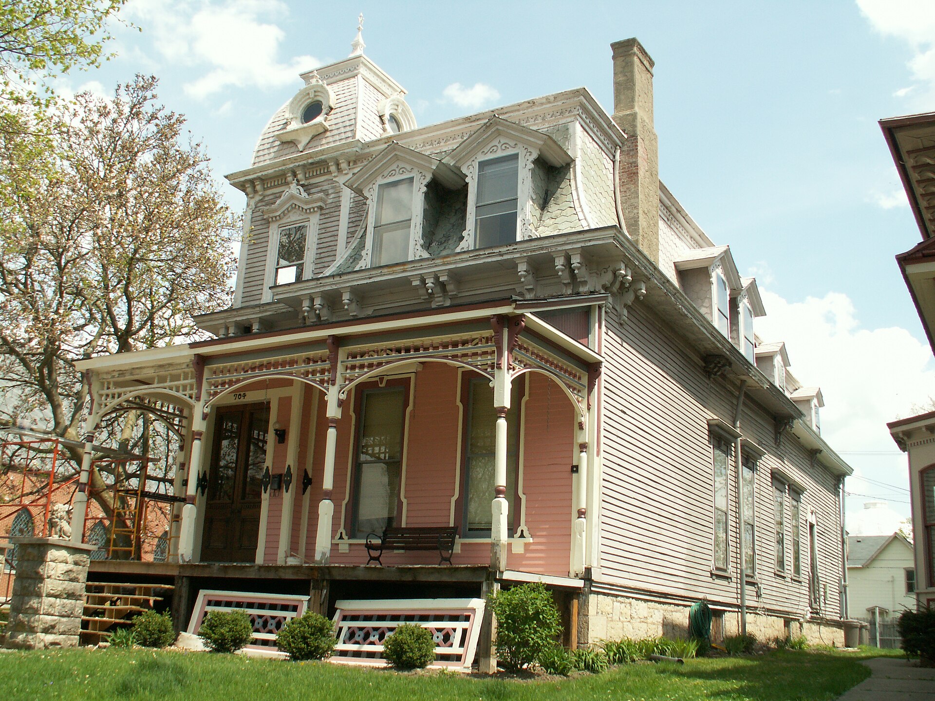 A tall wood-frame house with a mansard roof, a pink-and-white front porch, an empty bench on the porch and the number 704 over the steps, nobody in the frame