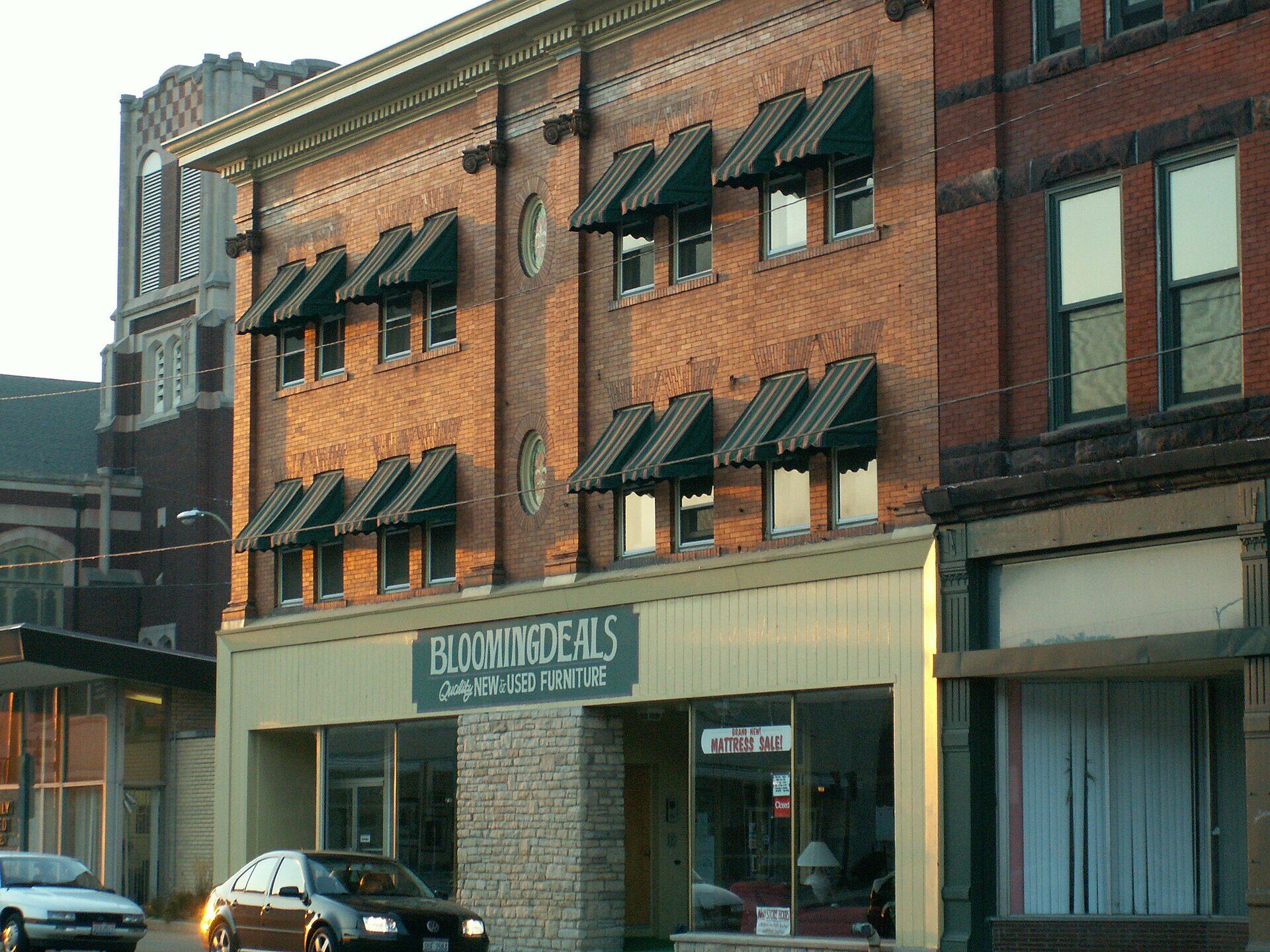 A three-storey red brick commercial building on a downtown street, with striped green awnings over the upper windows. The ground floor has a wide shop window under a painted sign reading BLOOMINGDEALS, Quality New and Used Furniture. A smaller sign in the window advertises a mattress sale, and a small red sign on the door reads Closed. Two parked cars stand at the kerb. No people are in the frame.