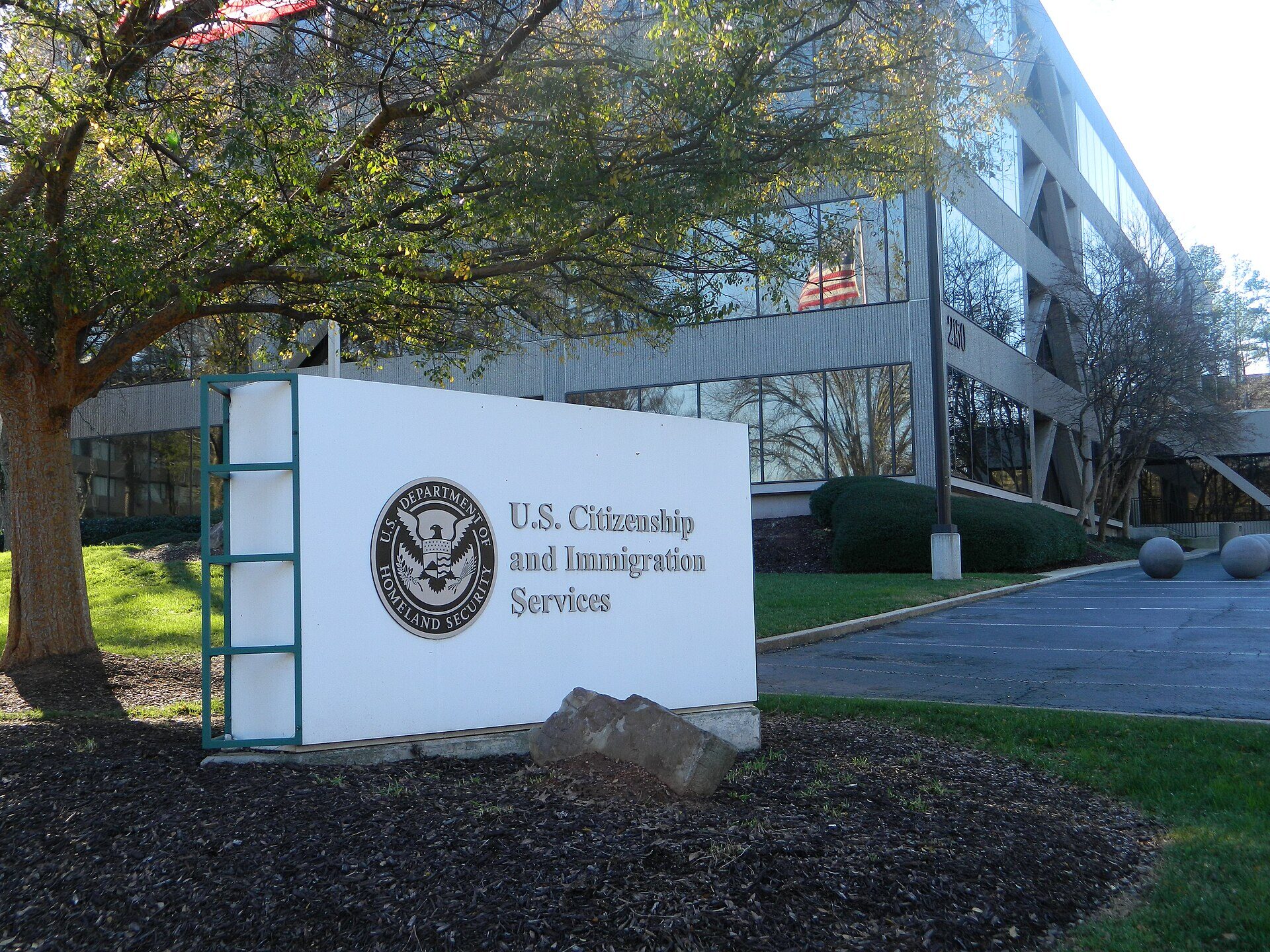 A low white monument sign in front of a glass and concrete office building. The sign carries the circular seal of the U.S. Department of Homeland Security and raised metal letters reading U.S. Citizenship and Immigration Services. A bare tree stands at the left, an American flag is reflected in the building's glass, and the car park beyond the sign is empty. No people are in the frame.